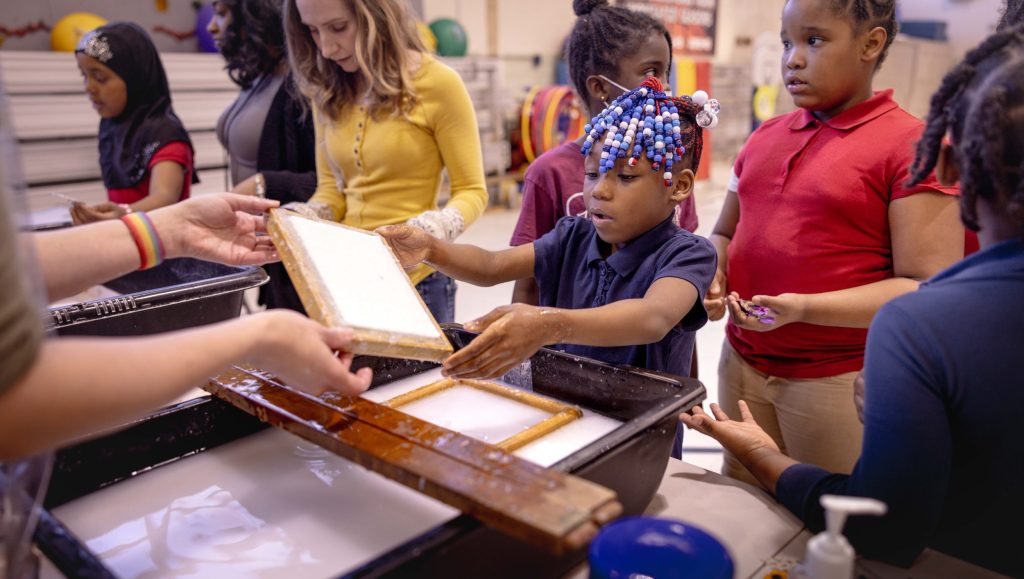 A group of children and adults watch a demonstration at a paper making station. One child helps to dip the paper frame into the bin of slurry.
