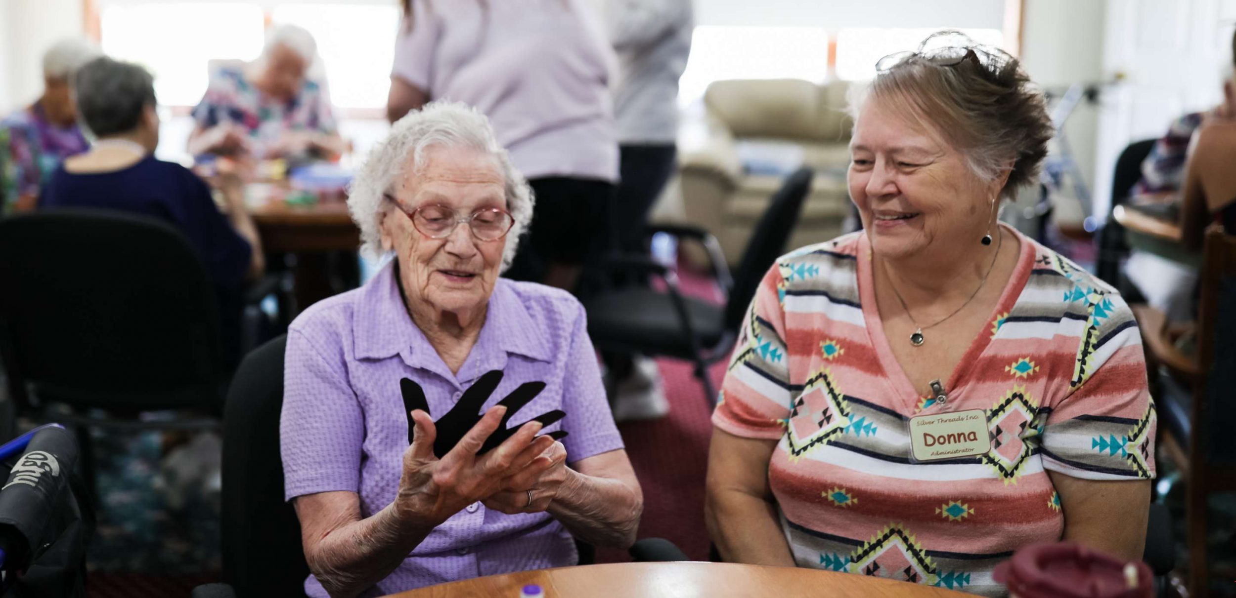 Two older light-skinned adults with short hair sit at a table together. One of the people is wearing a light purple shirt with curly short white hair and is holding a paper cut out of a hand up to their own hand and smiling.