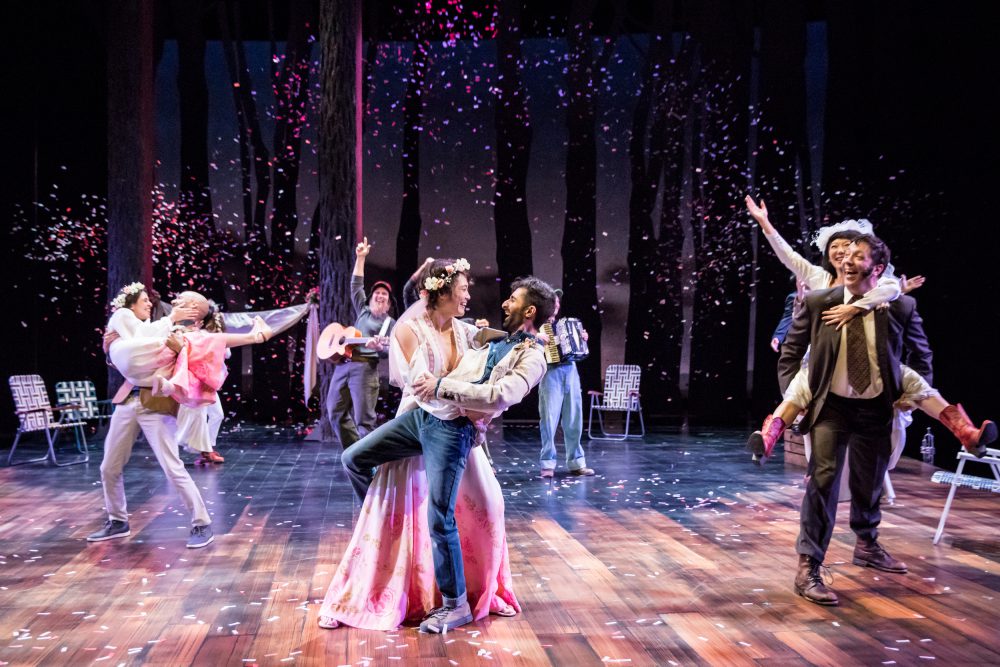 On a stage, couples are celebrating and holding each other. In the background, someone plays an acoustic guitar with a smile on their face and one arm reaching up, and another person plays an accordion.