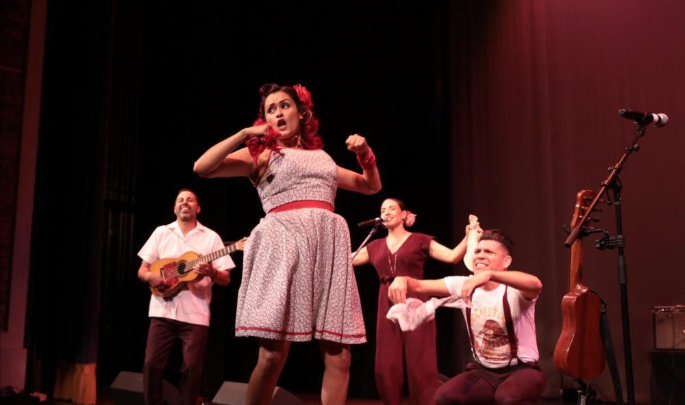 A group of four people performs on stage. The person in the front is turned to the side with their arms out in front of them like they are in the middle of a dance move.