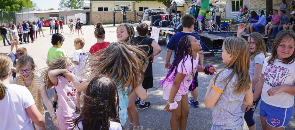 A group of children dance and laugh in a parking lot and a band performs behind them on a trailer.