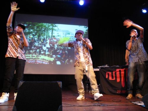 Three performers sing into mics while raising their hands upwards