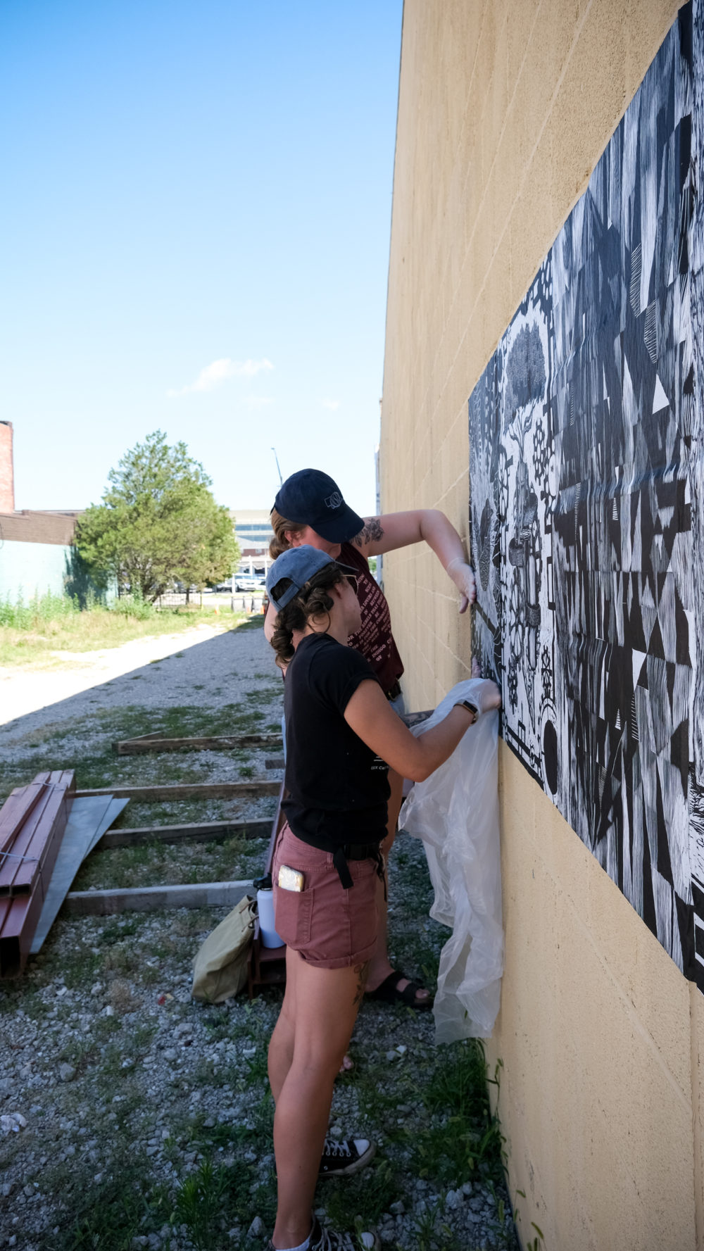 Two standing women wearing baseball caps paste a black and white print on a soft yellow wall