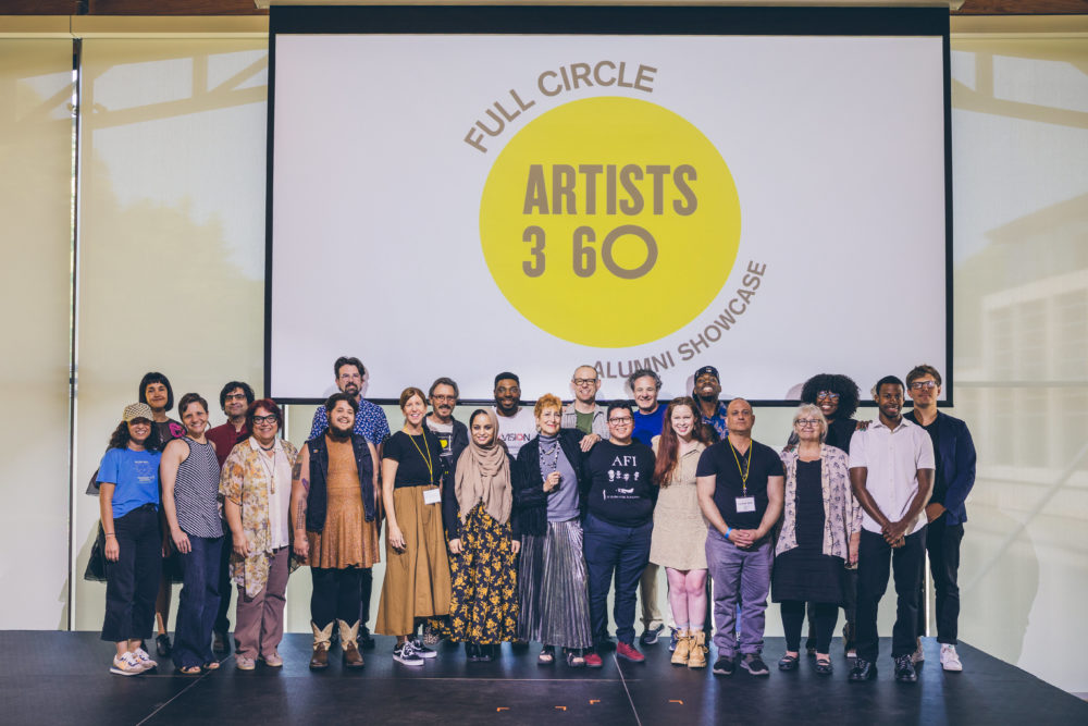 In front of a large screen that says Artists 360 Full Circle stand twenty artists in two rows