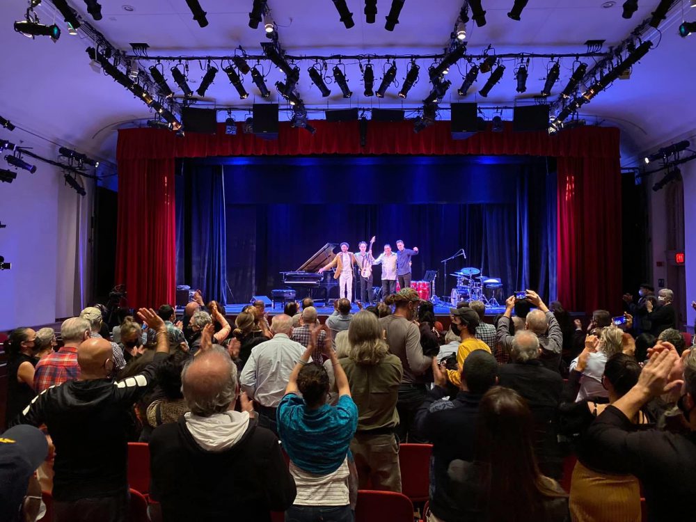 Shot from the back of the performance space, a group of four musicians stand on the stage in a row taking their final bow. They are surrounded by their instruments on a deep blue lit stage and framed with red curtains. The audience stands and claps.