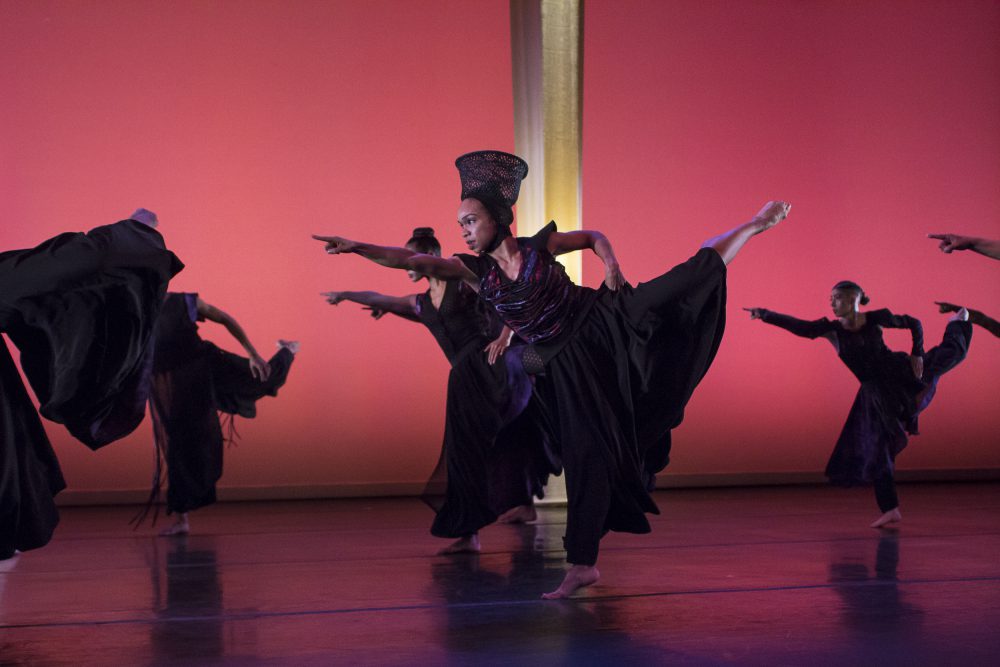 A group of seven dancers in elaborate outfits and headdresses extend their hands and legs upwards. They dance on an illuminated red stage.