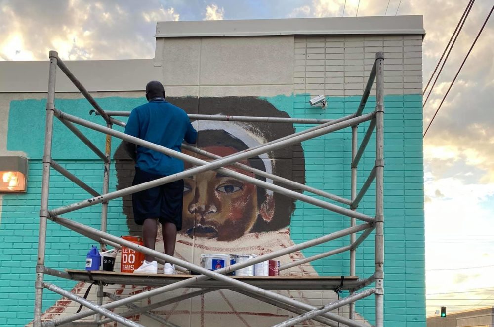 A man stand on a scaffold painting the head of an elaborate mural on a vibrant blue background.