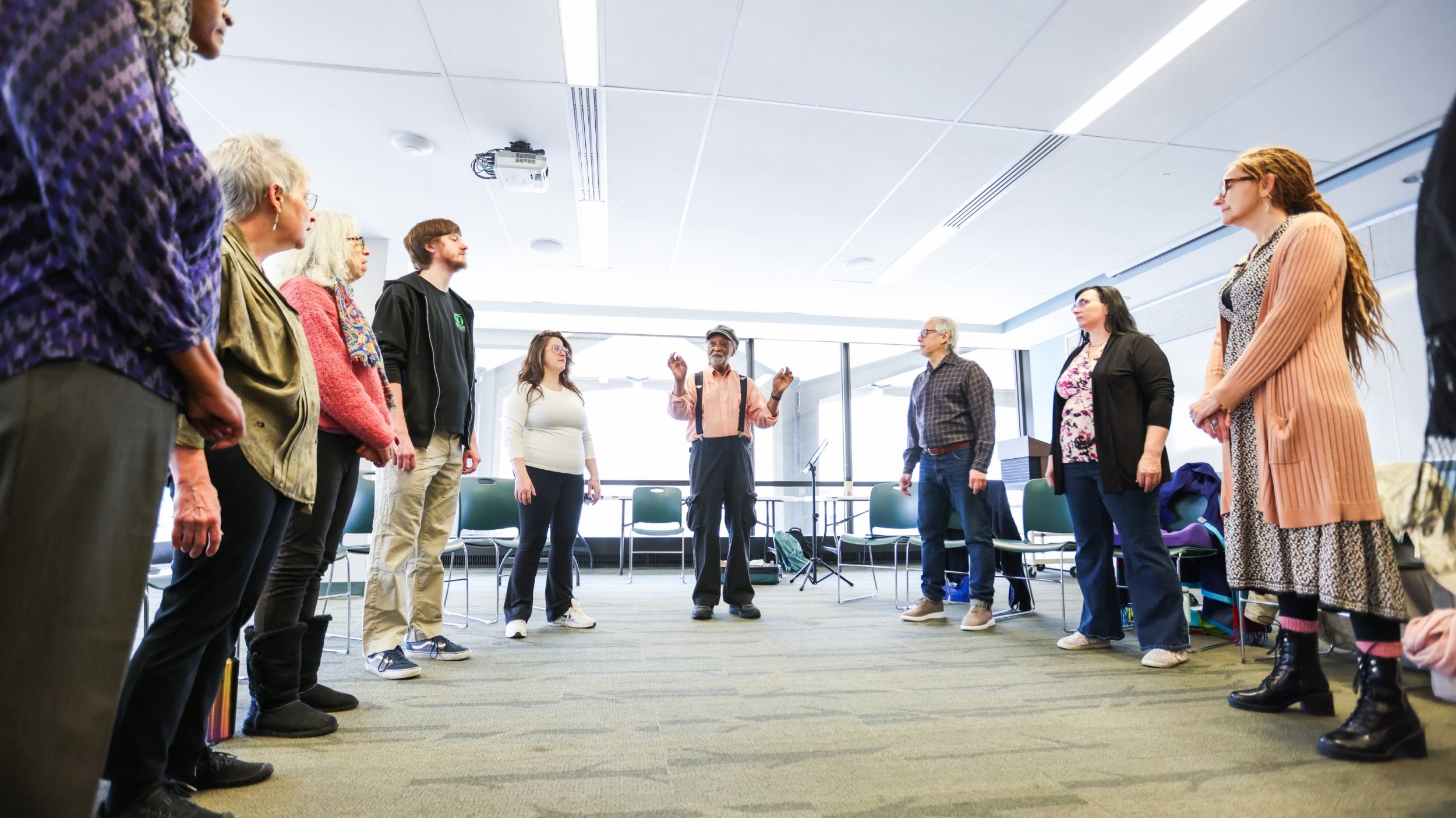 A group of adults stand in a circle in a bright meeting room, listening to a facilitator in the center who gestures as he speaks.